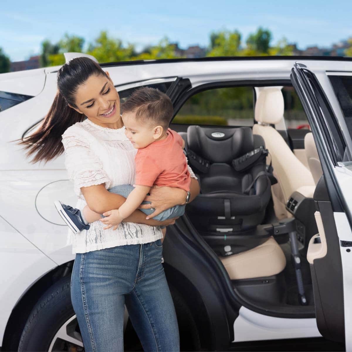 Mum holding little kid outside of the vehicle with Graco Turn2Me i-Size R129 rotating ISOFIX car seat. 